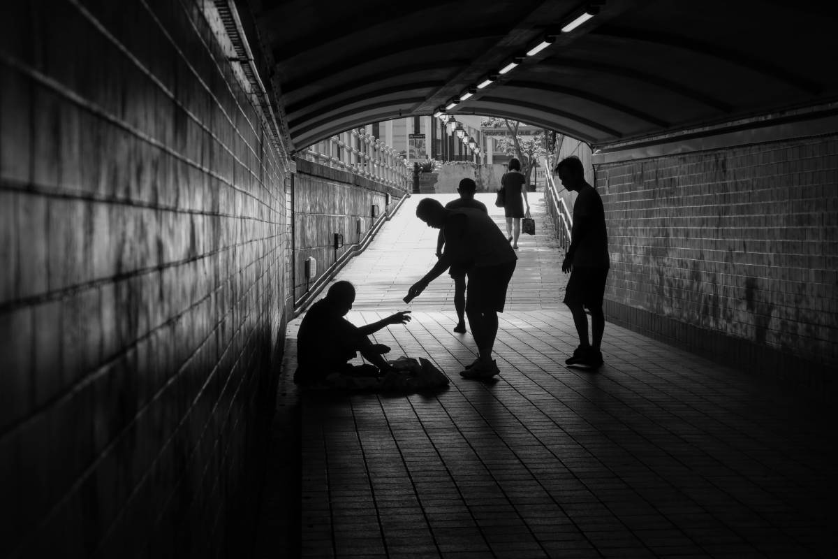 standing man giving sitting poor man in tunnel something in black and white photo